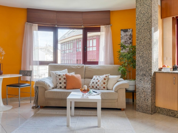 Sala de estar luminosa con un sofá en forma de L de color beige, una mesa de centro rectangular de madera con adornos y dos sillas tapizadas de color crema