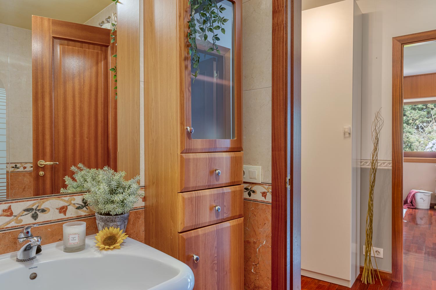 Cuarto de baño con mueble en madera y espejo decorado con elementos vegetales y puerta por que se ve otro dormitorio con ventana por la que se puede ver arboles.